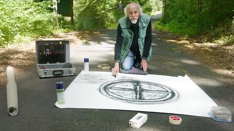 Der Künstler Benno Dalhoff sitzt auf dem Boden eines Waldweges vor einer Zeichnung, die er angefertigt hat. Neben ihm ein Koffer mit künstlerischen Werkzeugen.