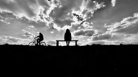 Motiv aus der Ausstellung Schonzone - Schwarz-Weiß-Aufnahme - eine Frau sitzt auf einer Bank - sie ist von hinten zu sehen, ein Fahrradfahrer fährt vorbei. Dramatischer Himmel.