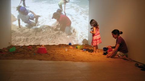 Kinder spielen im Sand, im Hintergrund auf einer Leinwand ebenfalls spielende Kinder.