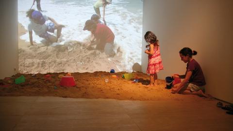 Kinder spielen im Sand, im Hintergrund auf einer Leinwand ebenfalls spielende Kinder.