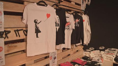 Blick in den Merch-Shop der Ausstellung "The Art of Banksy - Without Limits" in Frankfurt. Zu sehen sind T-Shirts mit Banksys Motiven.