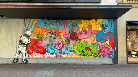 Das Bild zeigt ein buntes Graffiti an einer Wand in der Frankfurter Innenstadt. Darauf zu sehen ist ein Junge, der hinter einen Vorhang lugt. Hinter dem Vorhang sind bunte Smileys und andere Motive zu sehen.