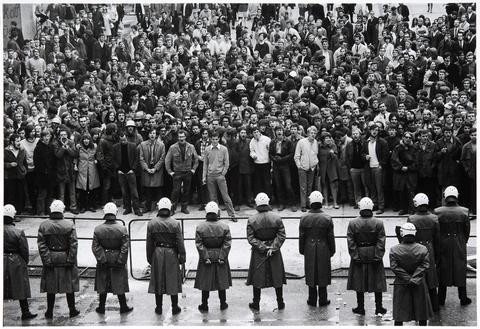 Schwarz-weiß-Fotografie: Eine große Gruppe von Menschen steht einer Reihe von behelmten Polizisten gegenüber