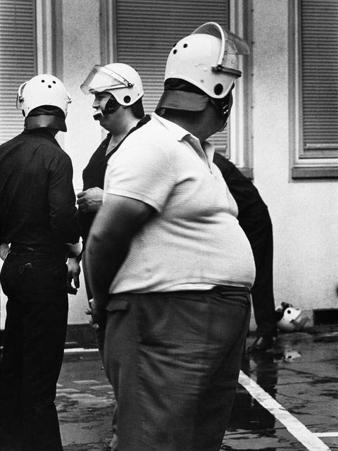 Ein sehr dicker Mann mit weißem T-Shirt und einem Helm, im Hintergrund weitere Männer mit Helmen