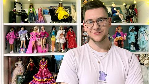 Ricardo Perez steht vor einem Regal, prall gefüllt mit Barbiepuppen, und lächelt in die Kamera.