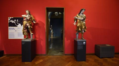 In einem Museumraum mit roter Wand stehen zwei fast mannshohe Engelsfiguren