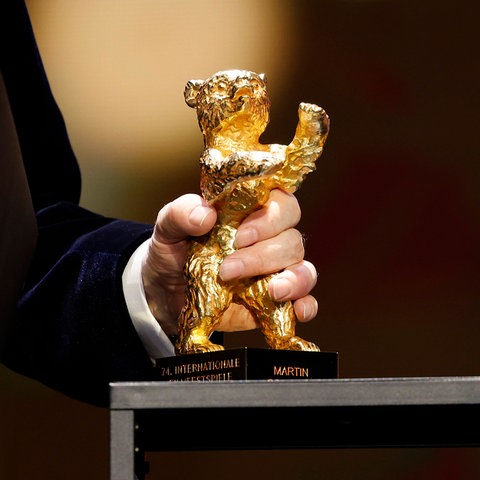 Das Bild zeigt die Auszeichnung Goldener Bär, der bei der Berlinale vergeben wird. Zu sehen ist eine Trophäe in der Form eines goldenen Bären. Der Sockel ist schwarz. Der Preis wird von einer männlichen Hand gehalten, zu sehen ist ein schwarzer Smoking-Ärmel.