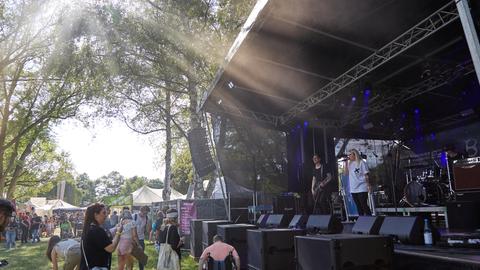Foto vom Besser-als-nix-Festival in Geisenheim aus dem Jahr 2019 - eine Musikbühne, davor vereinzelt Menschen, ein Mann im Rollstuhl. Auf der Bühne singt eine Frau, ein Mann spielt Gitarre. Die Bühne steht unter Bäumen. Im Hintergrund sind Pavillons zu sehen. 