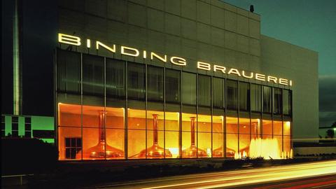 Das Bild zeigt das Binding-Sudhaus. Zu sehen ist ein rechteckiges Gebäude bei Nacht mit großer Fensterfront. Dahinter sind fünf große Kupferkessel zu sehen. Über dem Gebäude prangt der Schriftzug "Binding Brauerei".