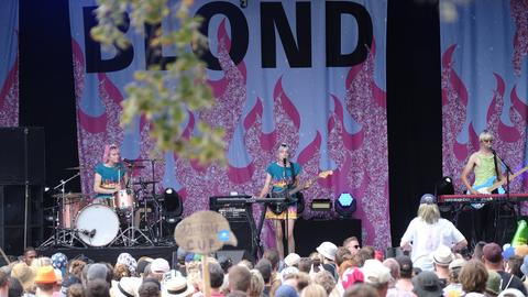 Drei Musikerinnen auf einer Bühne, im Hintergrund ein Banner mit dem Schriftzug "Blond".