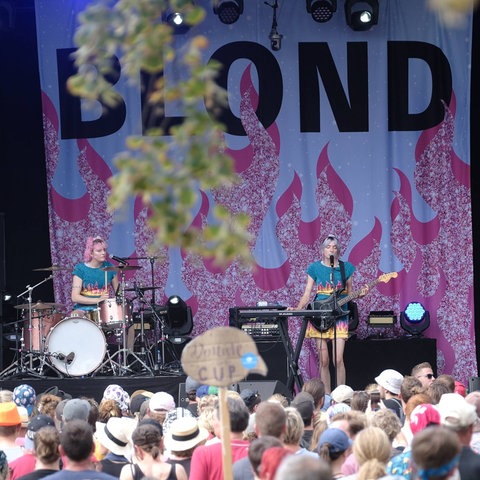 Drei Musikerinnen auf einer Bühne, im Hintergrund ein Banner mit dem Schriftzug "Blond".