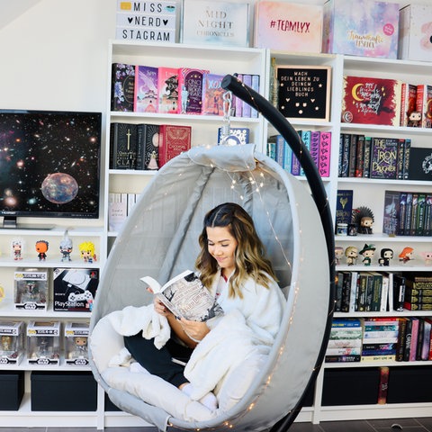 Junge Frau sitzt in Hängestuhl vor einem bunten Bücherregal und einem Bildschirm und liest