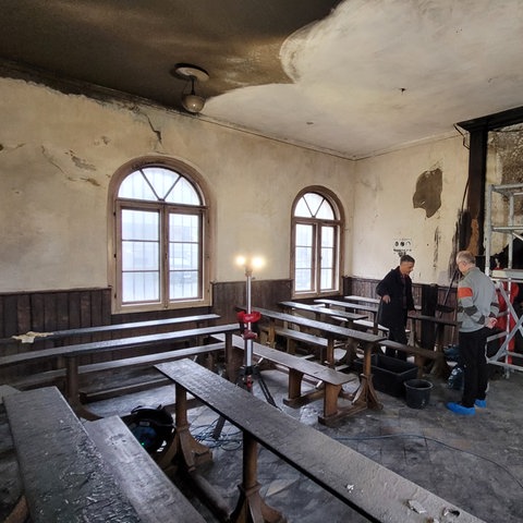Zwei Männer in einem russgeschwärzten, alten Gebäude, verkokelte Tische und Bänke