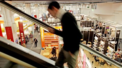 Eine Person fährt auf einer Rolltreppe hoch - sie ist nur verschwommen zu sehen - dahinter Blick auf eine Messehalle, in der nur wenige Menschen unterwegs sind. 