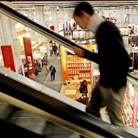 Eine Person fährt auf einer Rolltreppe hoch - sie ist nur verschwommen zu sehen - dahinter Blick auf eine Messehalle, in der nur wenige Menschen unterwegs sind. 