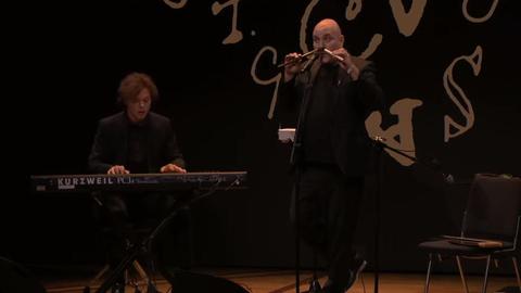 Zwei Männer in schwarzer Kleidung stehen während der Eröffnungsfeier der Buchmesse auf der Bühne. Links im Bild ist ein Mann mit halblangen braunen Haaren zu sehen, der Keyboard spielt, rechts neben ihm steht ein Mann mit Glatze, der zwei Flöten gleichzeitig spielt: mit dem Mund und der Nase.