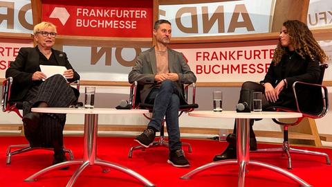 Das Bild zeigt zwei Frauen und einen Mann bei einer Diskussionsrunde auf der Frankfurter Buchmesse. Sie sitzen auf Stühlen auf einer Bühne, vor ihnen stehen zwei runde Tische mit Wassergläsern.