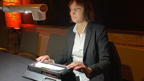 Eine Frau mit brauner Pagenfrisur sitzt an einem Tisch. Vor ihr steht ein Brailleschrift-Gerät, das wie eine Mischung aus Computer und Schreibmaschine aussieht.