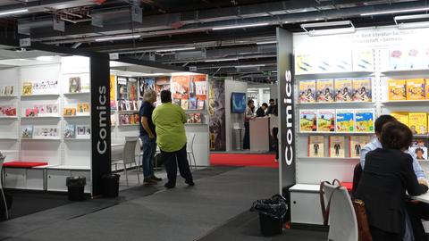 Das Bild zeigt einen Einblick in das Comics Centre auf der Frankfurter Buchmesse 2023. Links und rechts im Bild sind Kabinen verschiedener Verlage zu sehen. Links stehen zwei Männer mit dem Rücken zur Kamera und unterhalten sich, rechts sitzen zwei Menschen sitzen an einem Tisch. In den Regalen an den Wänden sind Comics aufgebaut.