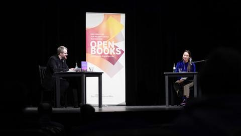 Ein Mann und eine Frau sitzen auf einer Bühne an Tischen mit Mikro und Wassergläsern. Daneben ein Aufsteller mit dem Logo von Open Books 2020. 