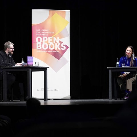 Ein Mann und eine Frau sitzen auf einer Bühne an Tischen mit Mikro und Wassergläsern. Daneben ein Aufsteller mit dem Logo von Open Books 2020. 