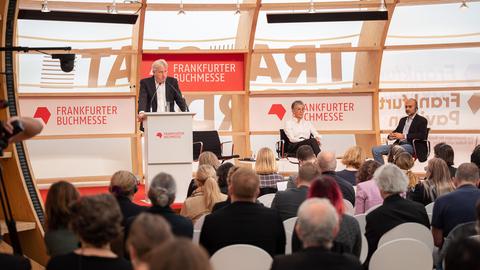Juergen Boos (links), Direktor der Frankfurter Buchmesse, spricht während der Eröffnungspressekonferenz. Karin Schmidt-Friderichs, Vorsteherin des Börsenvereins des Deutschen Buchhandels, und Schriftsteller Mohsin Hamid sitzen auf der Bühne.