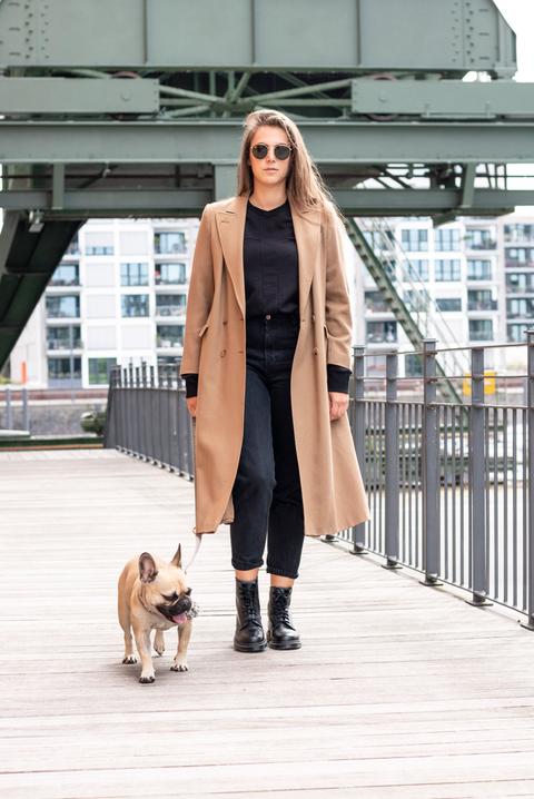 Eine Frau mit langem Camel-farbenem Mantel geht mit ihrem Hund spazieren