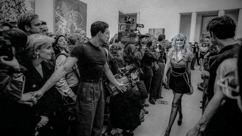 Claudia Schiffer schreitet durch eine Menge von Menschen, einige machen ihr den Weg frei, andere fotografieren oder filmen sie. Das Foto ist schwarz-weiß.