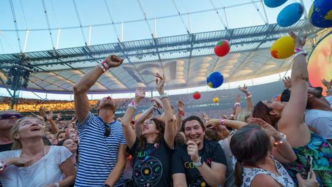 Fans in bunten Bandshirts stehen im Deutsche Bank Park beim Coldplaykonzert in der ersten Reihe.