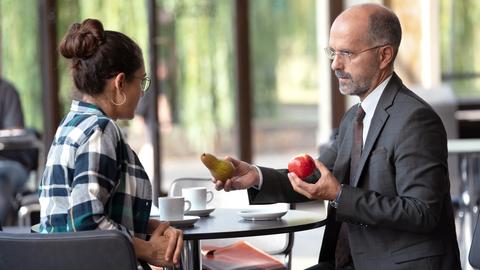 Filmszene aus "Contra": Ein Mann mit Glatze, Brille und Anzug und eine Frau mit dunklen, zu einem Knoten gebundenen Haaren und einem karierten Hemd sitzen sich an einem Bistrotisch gegenüber. Er hält ihr einen Apfel und eine Birne hin.