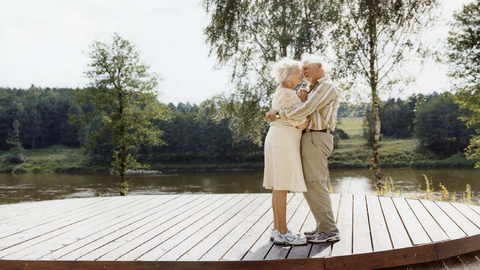 Ein älteres Paar hält sich umarmt. Es steht auf einen Holzbohlenuntergrund an einem Fluss. 