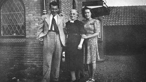 Das Schwarz-Weiß-Bild zeigt einen Mann, eine ältere und eine junge Frau vor einem Haus. Sie lächeln in die Kamera.