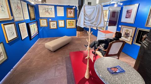 Ausstellungs-Ansicht der Schau "Dali" in Steinau an der Straße. Das Bild zeigt einen Raum mit blau gestrichenen Wänden, an denen viele gerahmte Gemälde hängen. In der Mitte des Raumes stehen ein Sessel, ein Garderobenständer aus Holz und ein Couchtisch auf einem roten Podest. In dem Sessel sitzt eine lebensgroße Silikon-Figur von Salvador Dali.