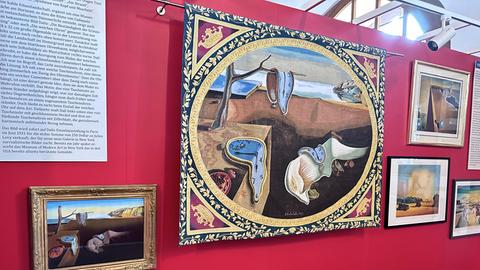 Ausstellungs-Ansicht der Schau "Dali" in Steinau an der Straße. Das Bild zeigt eine rot gestrichene Wand, an der gerahmte Gemälde hängen, darunter ein gold gerahmtes mit schmelzenden Uhren, die von einem Tisch herunterlaufen.