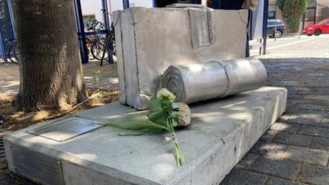 Das Bild zeigt eine Betonskulptur in der Form eines Koffers. Er ist auf einer Betonplatte aufgestellt, davor liegen zwei Rosen.