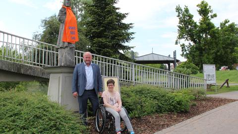 Ein orangeroter Schal mit der Aufschrift "Welt-MS Tag" schmückt die Nepomuk-Statue neben der Fußgängerbrücke über den Altrhein in Riedstadt-Erfelden. Auch auf dem Bild: Bürgermeister Marcus Kretschmann und die Behindertenbeauftragte Marina Wohner im Rollstuhl