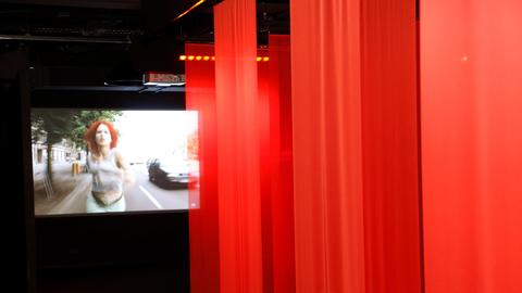Das Bild zeigt eine Leinwand, auf der eine Szene aus dem Film "Lola rennt" zu sehen ist. Am rechten Bildrand sind rot beleuchtete Vorhänge zu sehen.