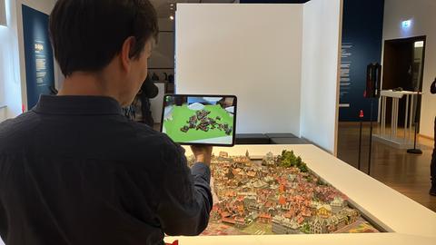 Mann hält Tablet über Modell einer Stadt. Auf dem Bildschirm erscheint ein gezeichnetes Bild der Stadt.