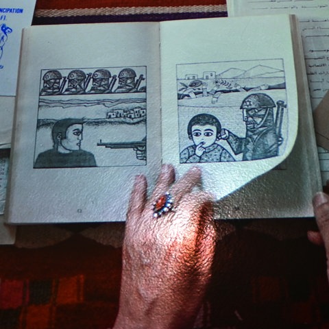 Screenshot einer Projektion: Eine Hand blättert in einem Heft mit einer Bilderzählung. Daneben liegen weitere Hefte, Bilder und Karten.