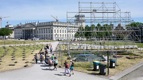 Eine Gruppe von Menschen läuft an dem Gerüst vorbei, an dem bis vor kurzem die umstrittene künstlerische Arbeit hing. Im Hintergund der Friedrichsplatz und das Fridericianum. (Vogelperpsektive)