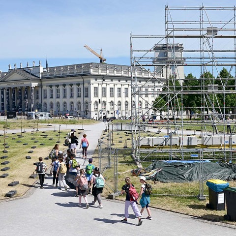 Eine Gruppe von Menschen läuft an dem Gerüst vorbei, an dem bis vor kurzem die umstrittene künstlerische Arbeit hing. Im Hintergund der Friedrichsplatz und das Fridericianum. (Vogelperpsektive)