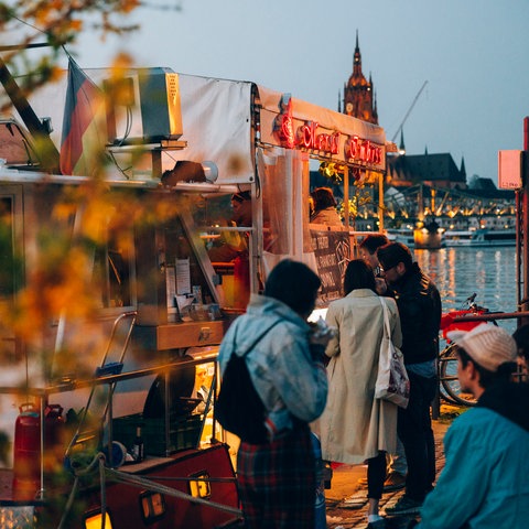 Leute stehen in der Abenddämmerung vor dem Dönerboot am Mainufer. Im Hintergrund der Frankfurter Dom.