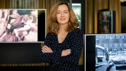 Das Bild zeigt Museumsdirektorin Ellen Harrington im Deutschen Filmmuseum. Sie trägt hellbraune, schulterlange Haare und ein blaues Kleid mit weißen Punkten. Sie lächelt in die Kamera. Hinter und neben ihr zeigen Bildschirme Filmszenen.
