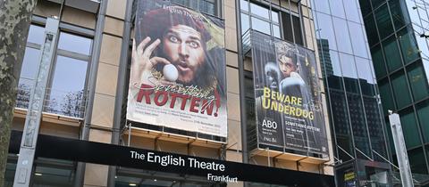 Auf einem Plakat über dem Eingang des Theaters wird das Musical "Something Rotten" beworben. 