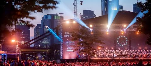 Im Dunkeln rot-lila beleuchtete Bühne mit Orchester und Publikum davor. Im Hintergrund die Frankfurter Skyline