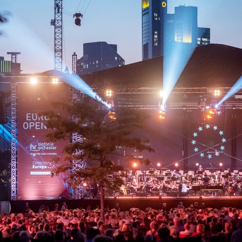 Im Dunkeln rot-lila beleuchtete Bühne mit Orchester und Publikum davor. Im Hintergrund die Frankfurter Skyline