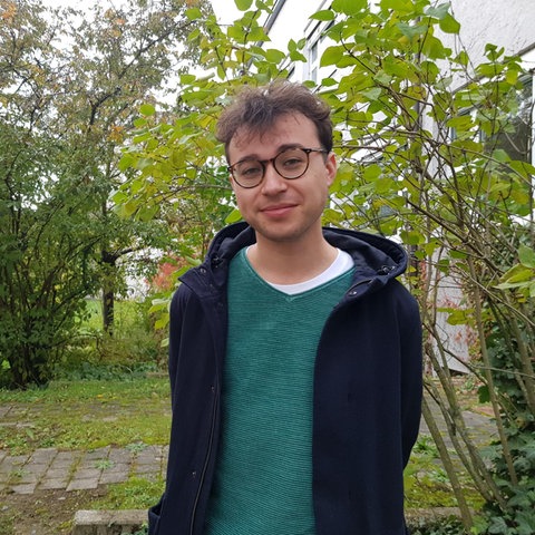 Saxofonist Fabian Dudek blickt in die Kamera. Hinter ihm sind Büsche und Sträucher zu sehen.