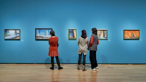 Drei Frauen stehen vor einer blauen Wand, an der mehrere Ölgemälde hängen.