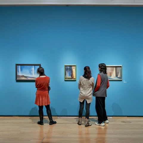 Drei Frauen stehen vor einer blauen Wand, an der mehrere Ölgemälde hängen.