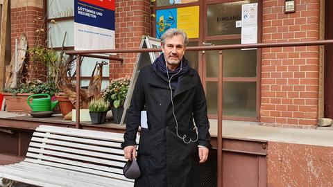 Ein Mann mit grauen Haaren, grauem Bart und schwarzer Jacke steht vor einem Backstein-Gebäude. Neben ihm ist eine Bank aus Holz. Er trägt weiße Kopfhörer, das Kabel hängt über der Jacke.